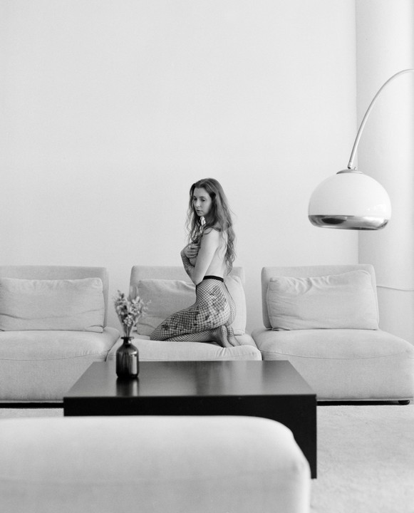 A woman kneels on a sofa in a living room, covering her chest with her arms.
