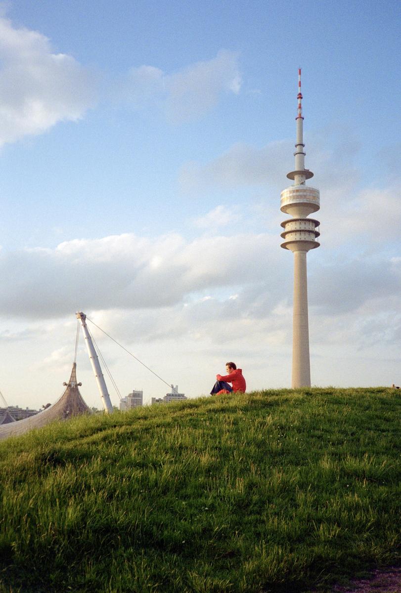 A man sits on a grassy hill with a tall telecommunications tower and a large tent-like structure in the background.