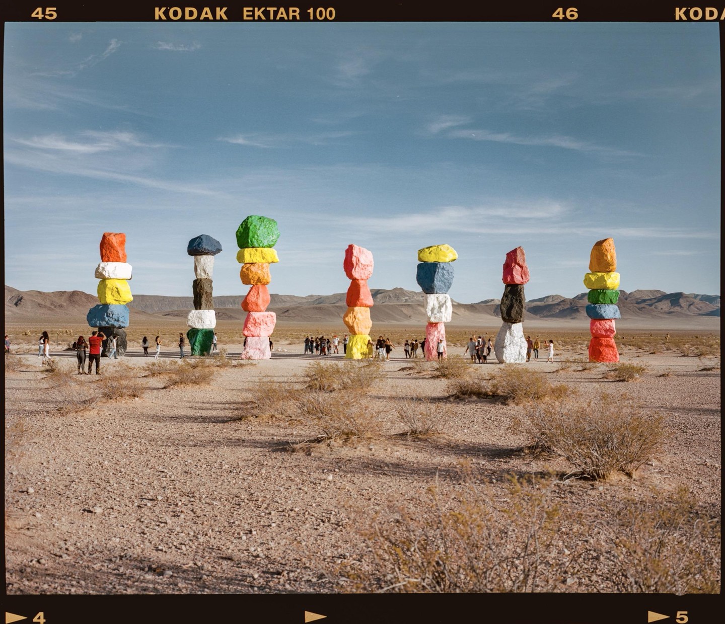 Seven stacks of brightly painted boulders stand in a desert landscape with mountains in the distance and people walking around them.