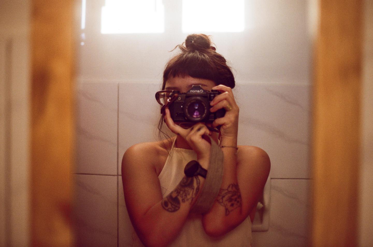 A woman with a bun and bangs takes a mirror selfie with a camera, revealing tattoos on her arms.