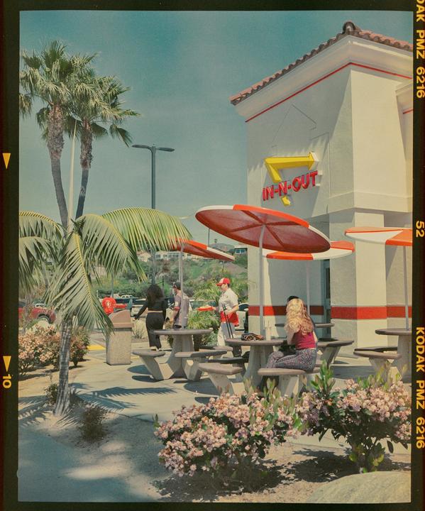 People are gathered outside an In-N-Out Burger restaurant with palm trees and flowering bushes.