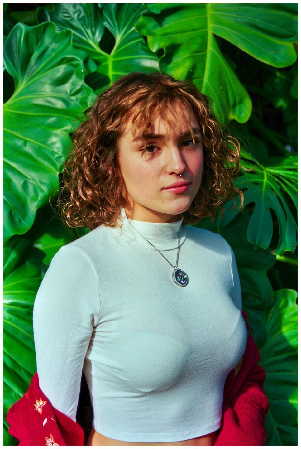 A woman with curly hair and a white turtleneck stands in front of large green leaves.