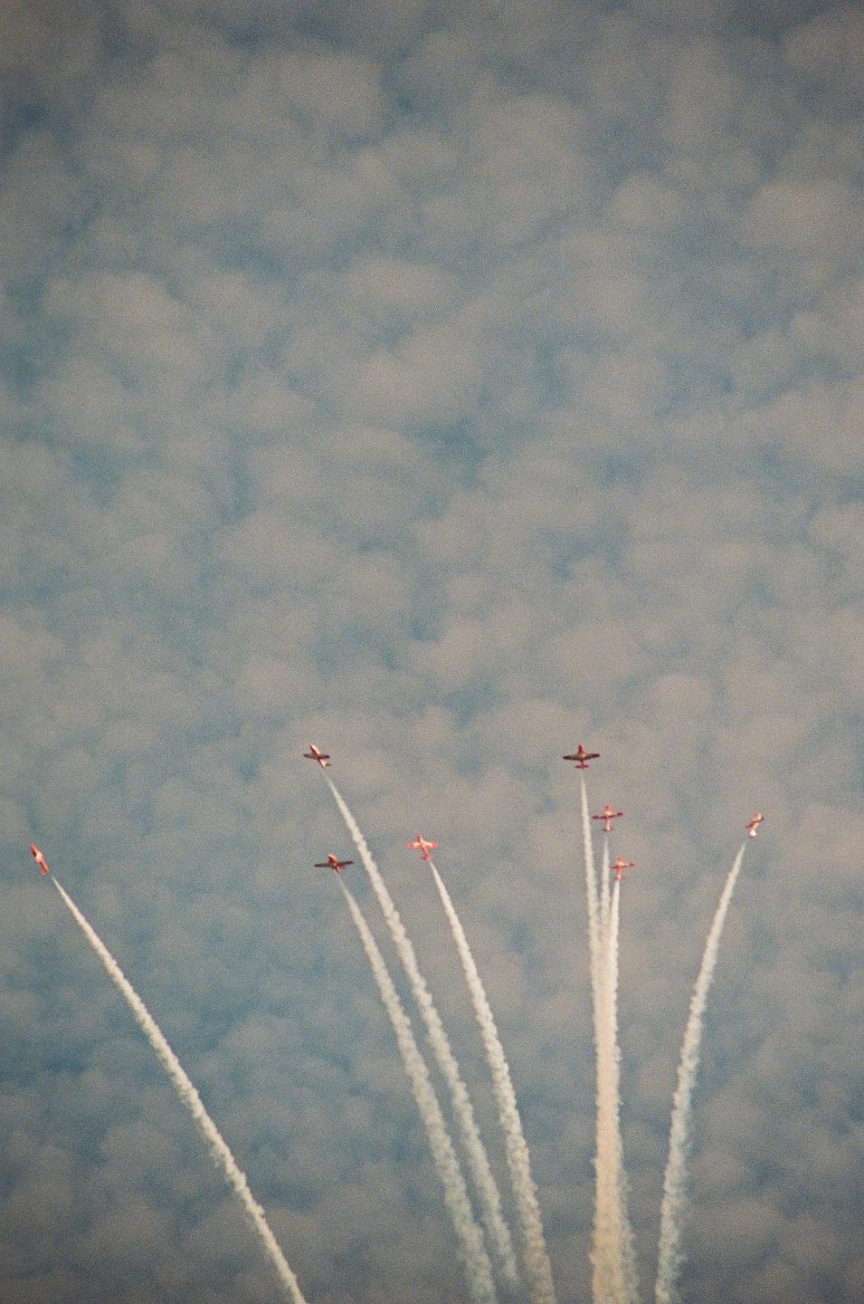 Several small airplanes fly in formation, leaving white contrails against a cloudy sky.