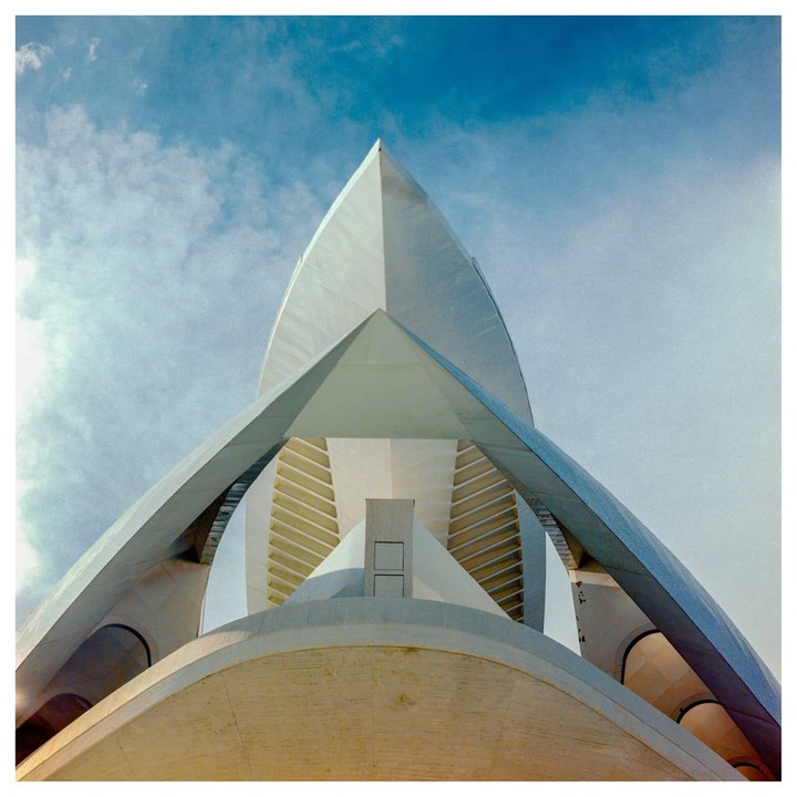 A low-angle view shows the top of a modern building with a pointed roof and curved architectural elements against a blue sky.