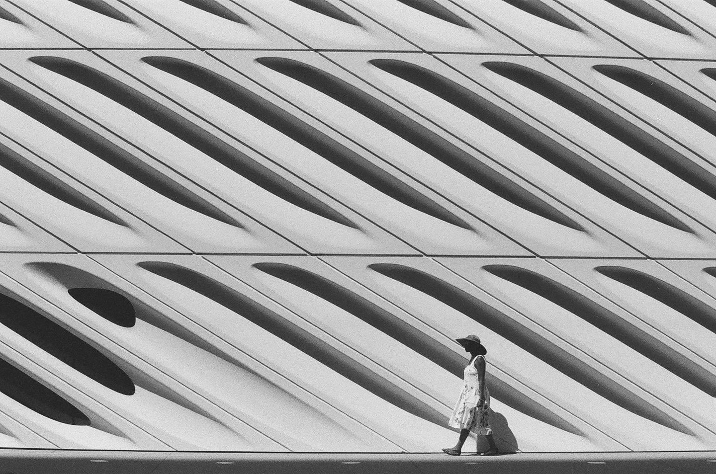 A woman in a hat and dress walks past a modern building with diagonal patterns and shadows.