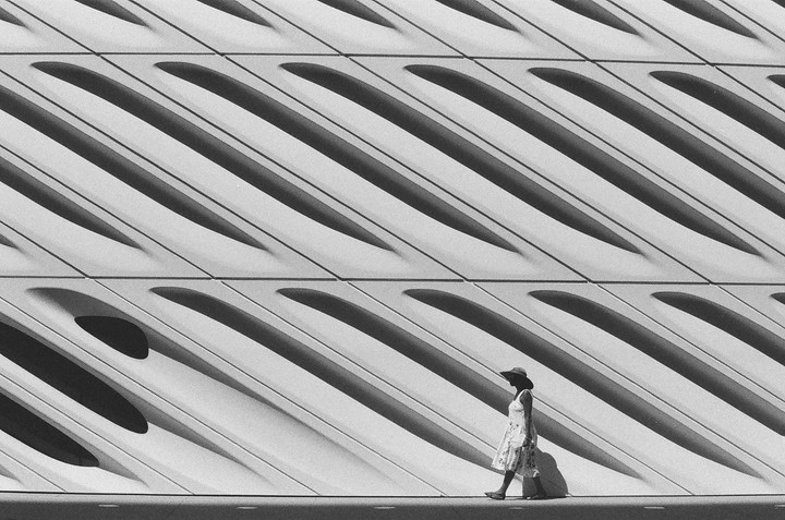 A woman in a hat and dress walks past a modern building with diagonal patterns and shadows.