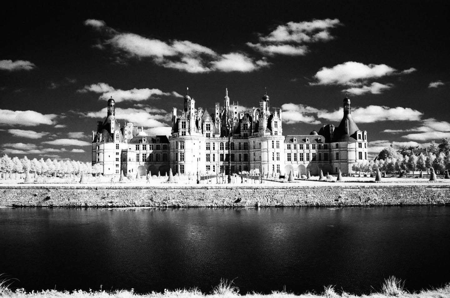 A large castle with many turrets and windows stands behind a stone wall and a body of water under a cloudy sky.