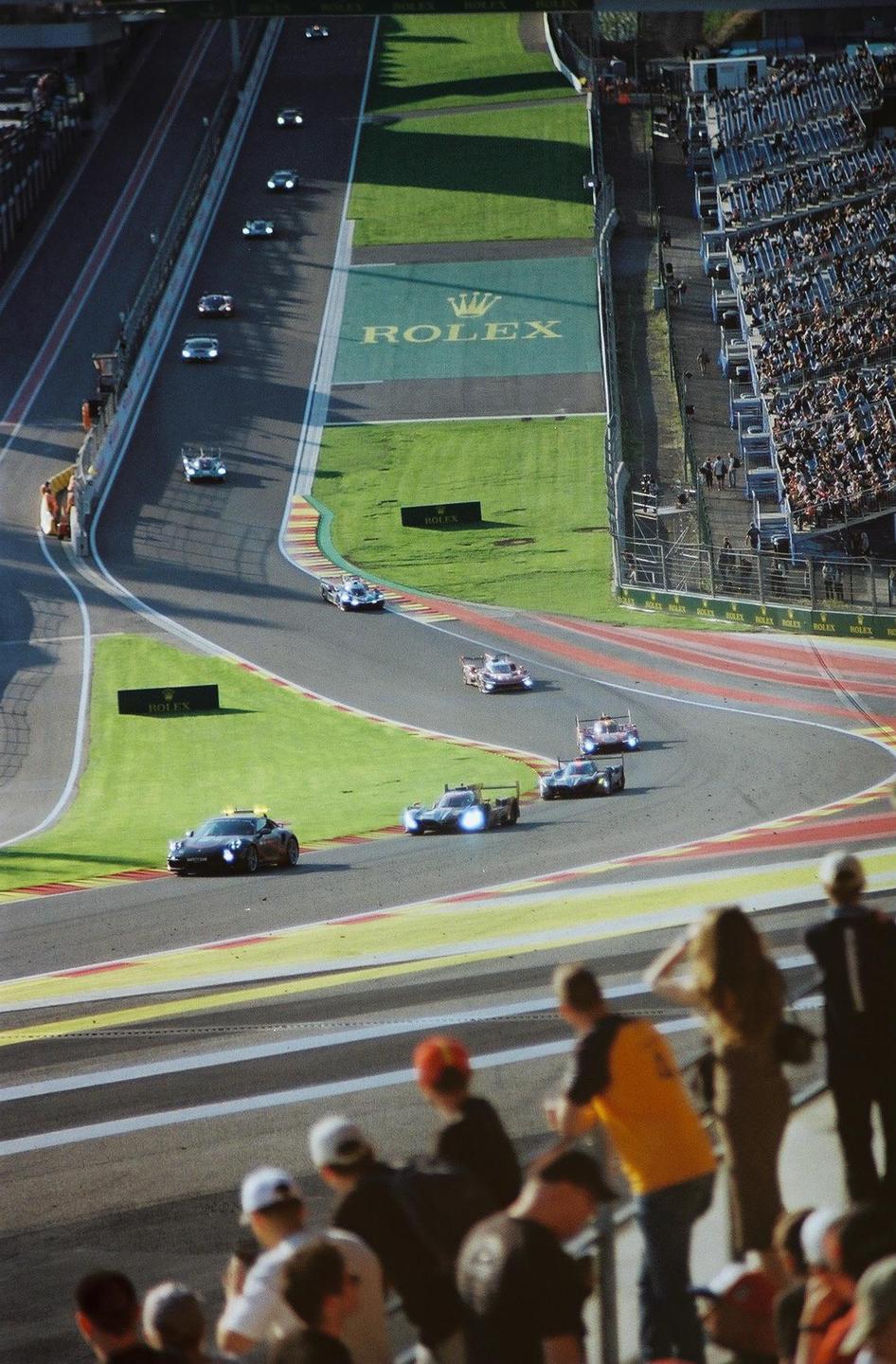 A race track with multiple cars and spectators in the stands is shown from an elevated perspective.