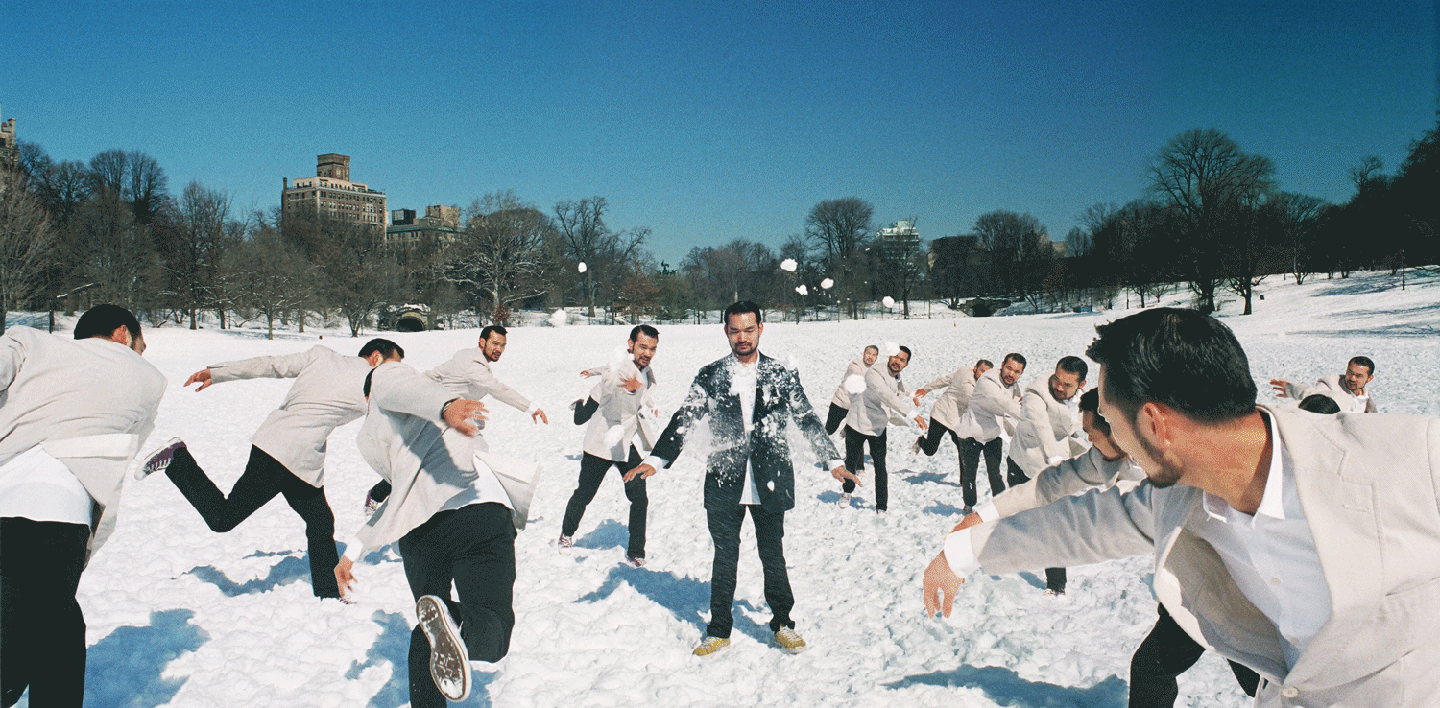 A man in a dark suit is pelted with snowballs by multiple versions of himself in a snowy park.