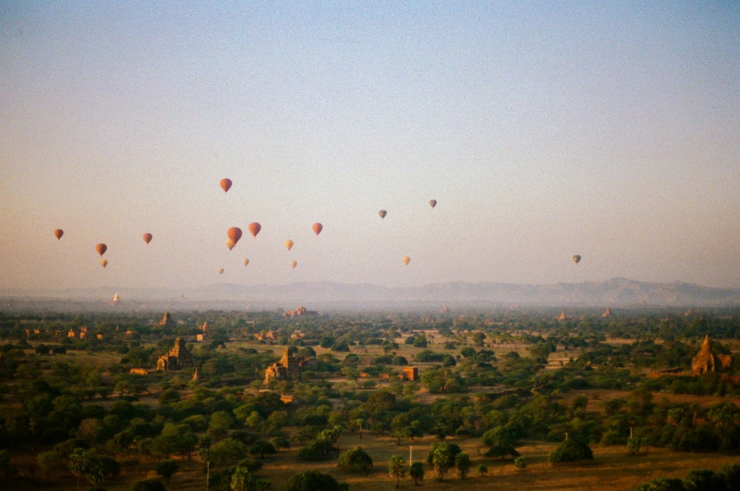 Hot air balloons float over a landscape filled with ancient stupas and trees at sunset.