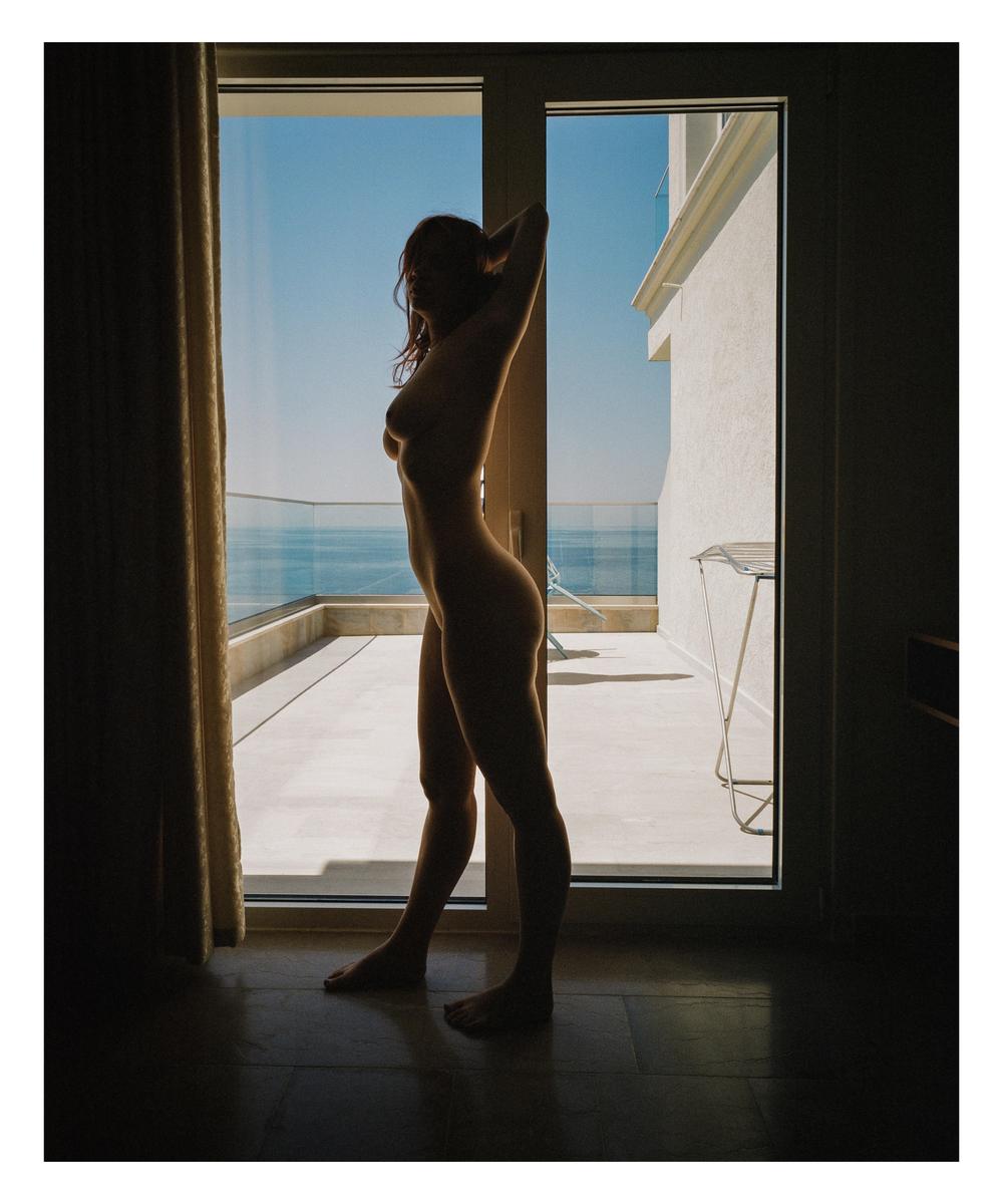 A nude woman stands in silhouette in a doorway looking out at the ocean from a balcony.