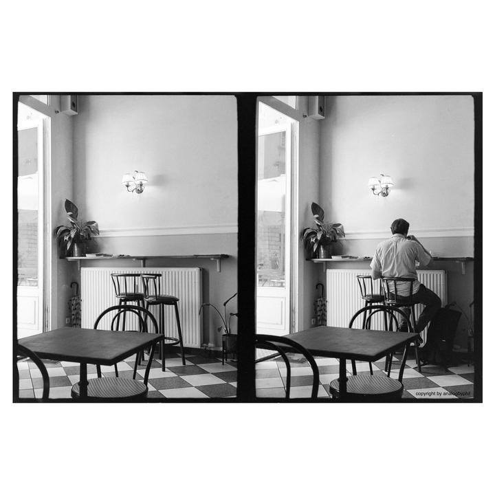 A man sits at a counter in a cafe with a checkered floor and potted plants.
