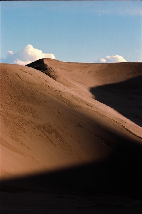 A large sand dune is partially covered in shadow under a blue sky with white clouds.