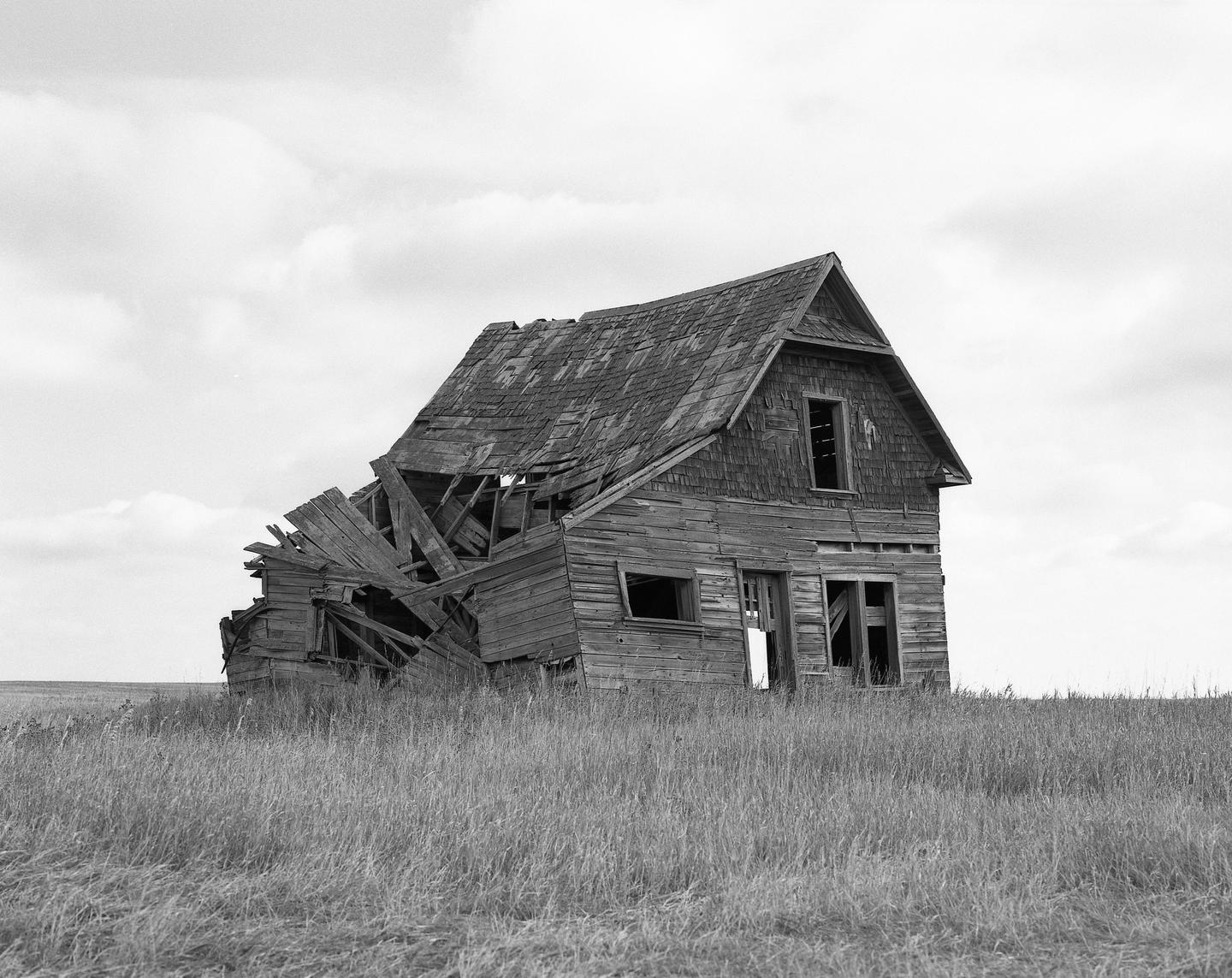 An abandoned wooden house with a collapsed side stands in a field of tall grass under a cloudy sky.