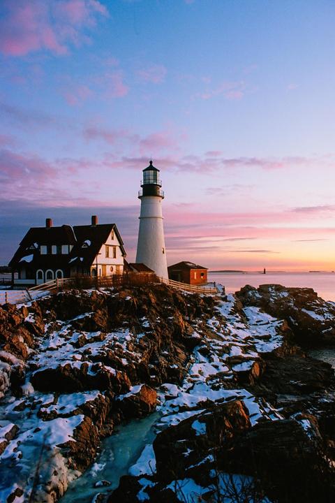 A lighthouse and a house stand on a rocky, snow-covered coast under a colorful sky at sunrise.
