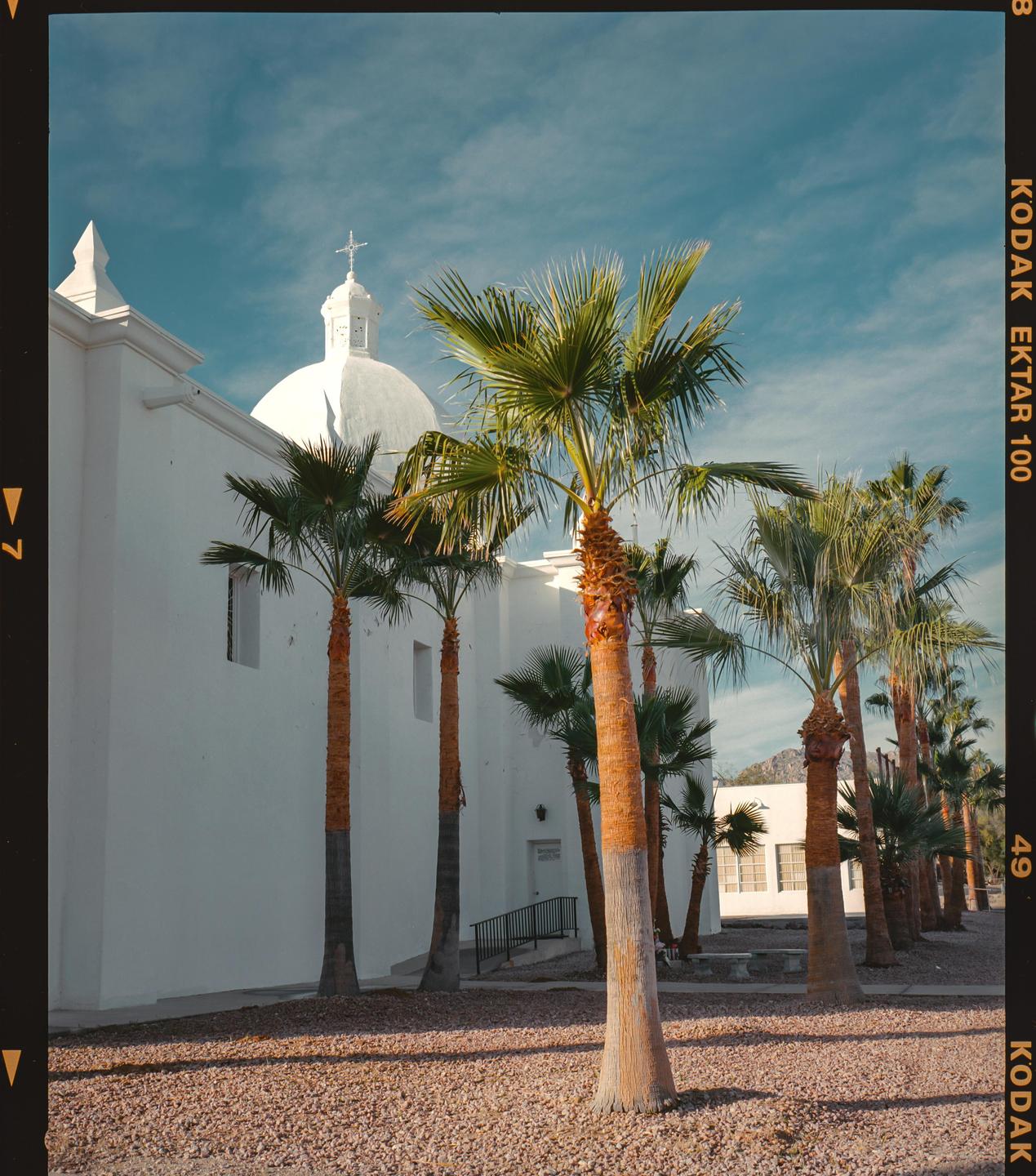 A white building with a dome and cross on top is surrounded by palm trees under a blue sky.