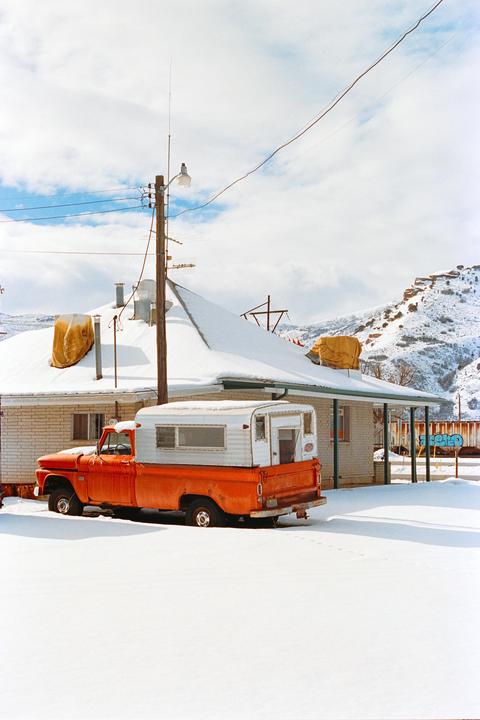 An orange pickup truck with a camper shell is parked in the snow next to a house with a snow-covered roof, with a mountain in the background.