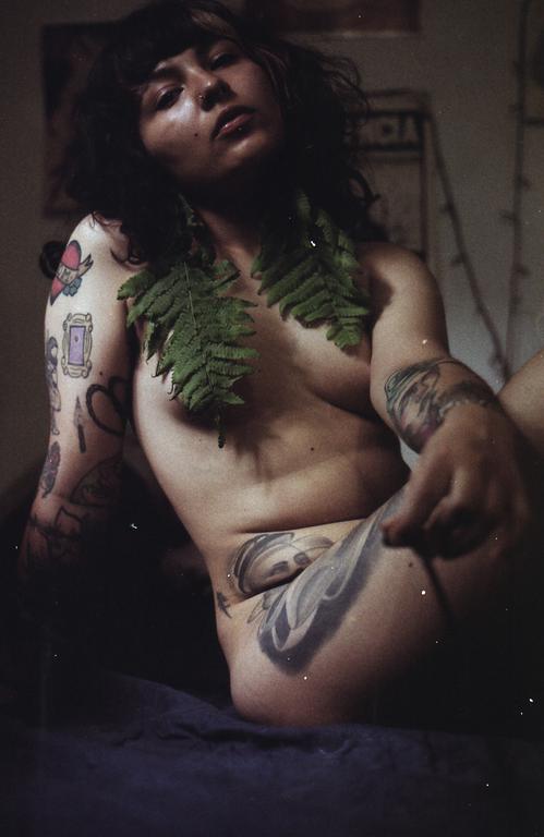 A woman with tattoos and fern leaves around her neck is reclining on a dark surface.