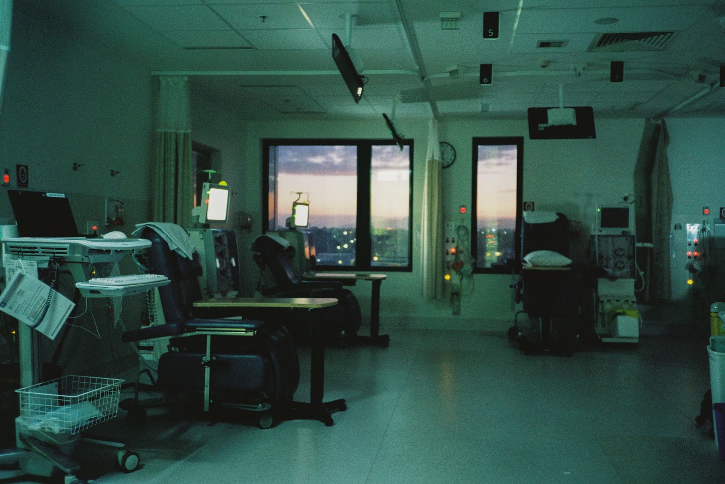 A hospital room with several medical chairs and equipment is illuminated by the early morning light filtering through large windows.