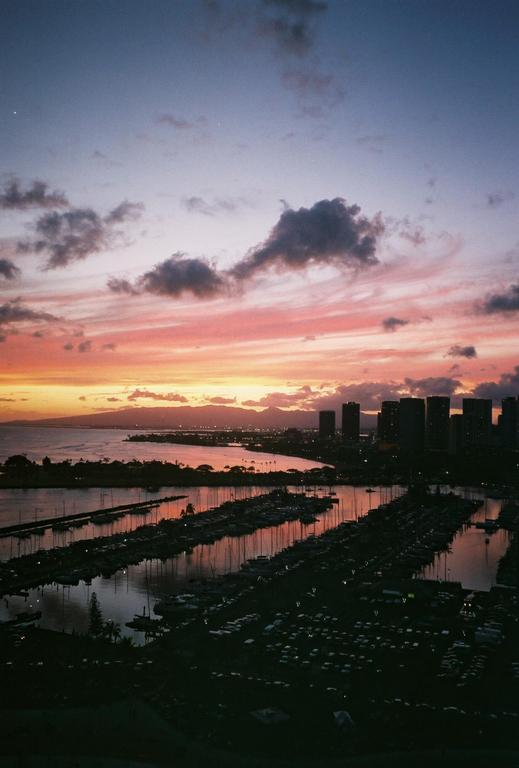 A vibrant sunset illuminates the sky above a marina filled with boats and a city skyline in the distance.