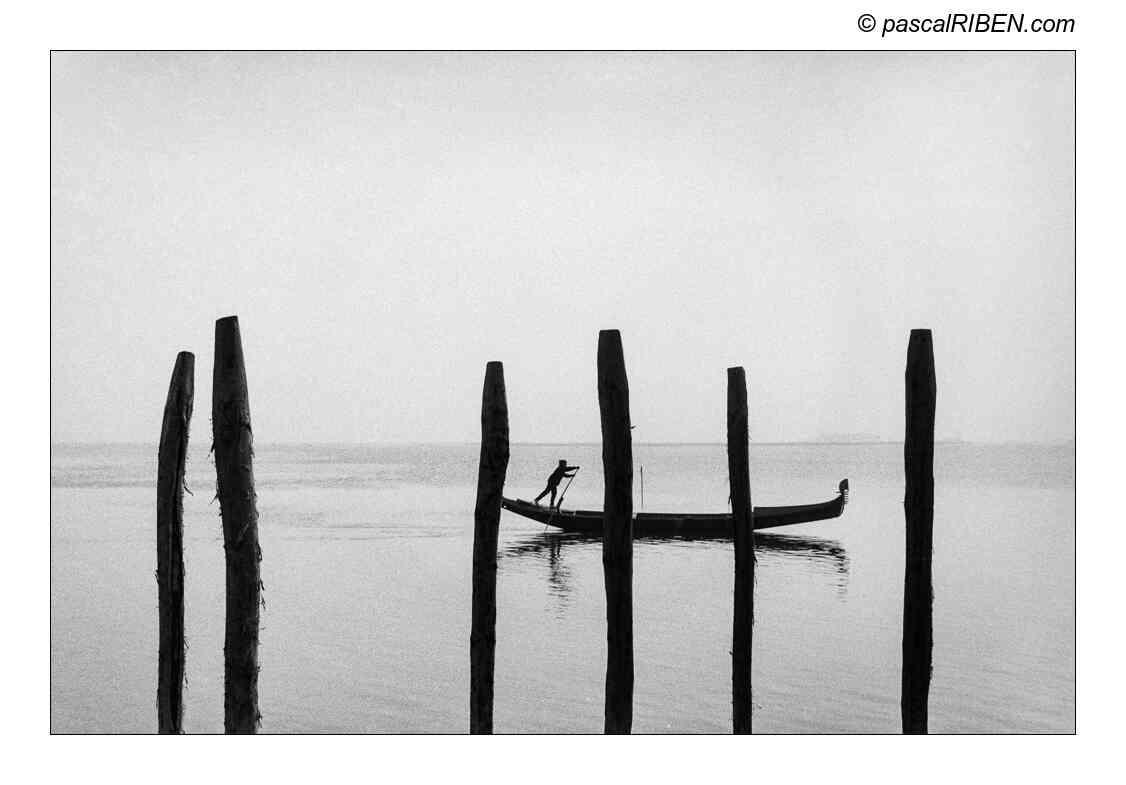 A gondolier poles a gondola between wooden posts in the water.