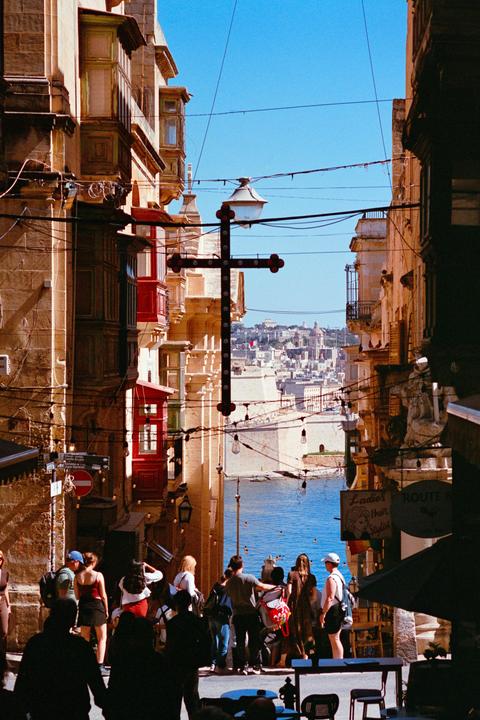 A street in Valletta is filled with people and buildings with colorful balconies, leading to a view of the sea and a distant city.