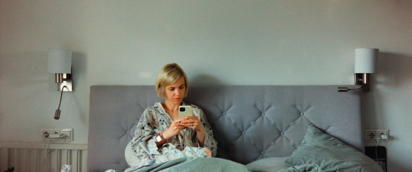 A woman sits in bed wearing a patterned robe and looking at her phone.