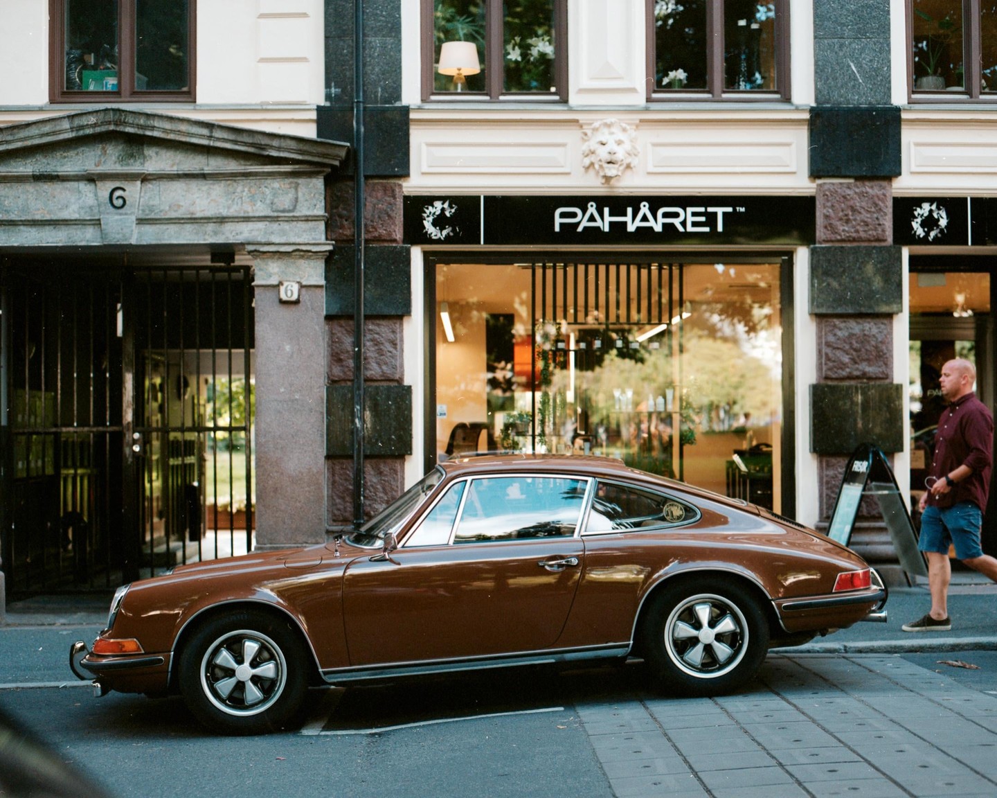A brown Porsche is parked on a city street in front of a building with a storefront and a man walks past it.