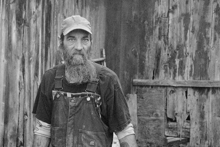 A man with a long beard and a baseball cap stands in front of a weathered wooden wall.