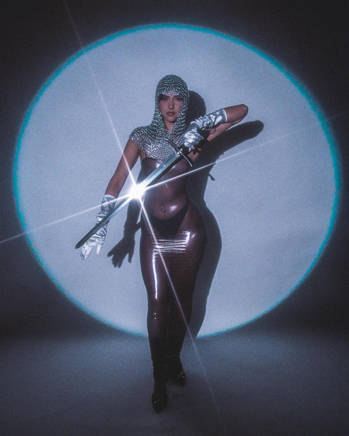 A woman in a chainmail coif and gloves holds a sword in front of a spotlight.
