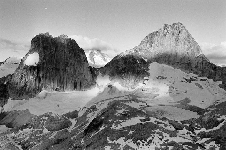 Two large, rugged mountain peaks with snow and glaciers are visible under a cloudy sky, with a small moon in the upper left.