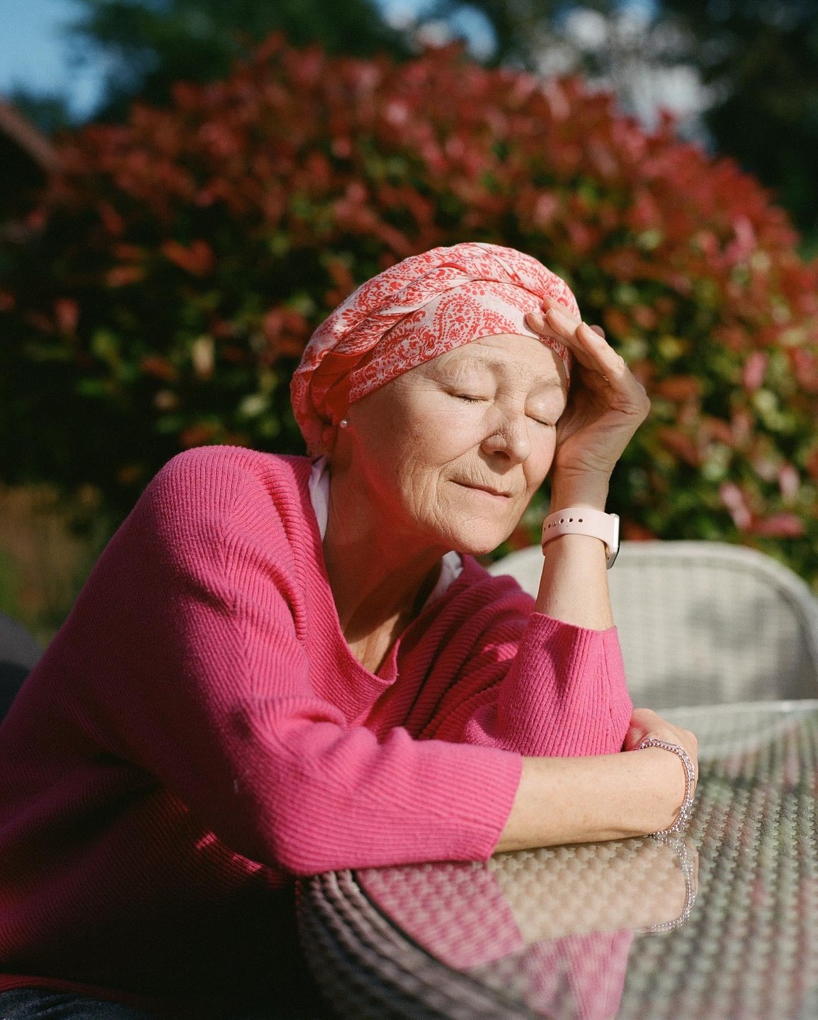 A woman with a headscarf sits at a table with her eyes closed and her hand on her forehead.