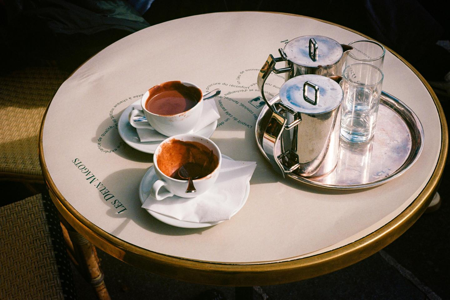 Two cups of hot chocolate and two metal pitchers sit on a round table at Les Deux Magots.