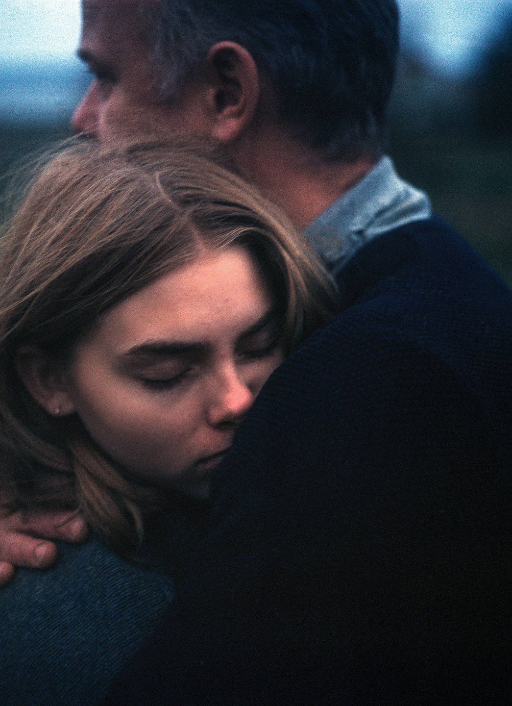 A young woman with closed eyes rests her head on a man's shoulder as they embrace.