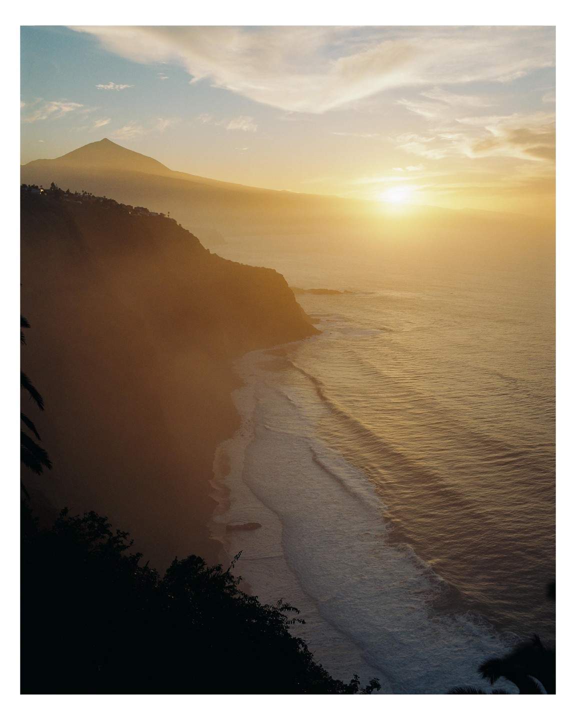 The sun sets over a mountain and ocean with waves crashing on a beach.