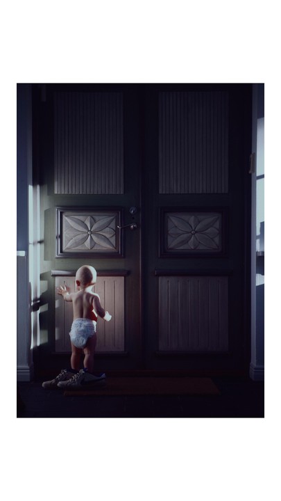 A baby in a diaper stands in front of a dark door with two pairs of shoes on the floor.