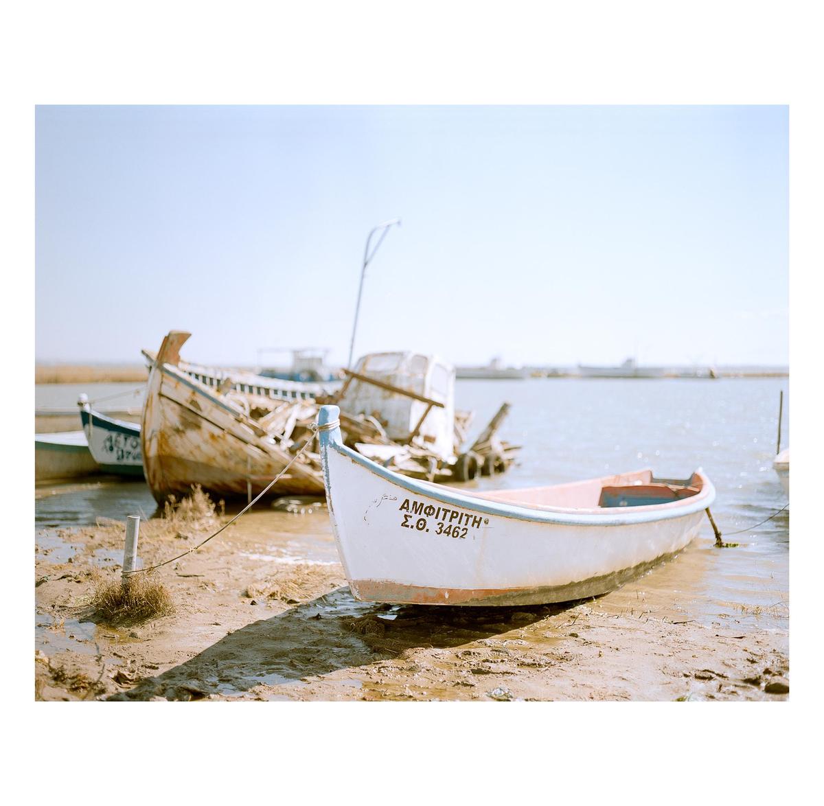 A small white boat with a blue trim is pulled ashore in the foreground, with a dilapidated wooden boat behind it and water in the background.