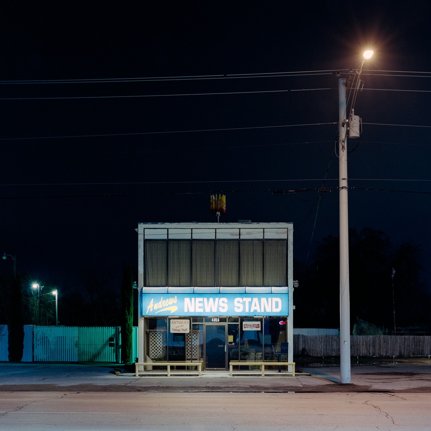 A news stand with a bright blue sign is illuminated at night, with a utility pole and power lines visible.