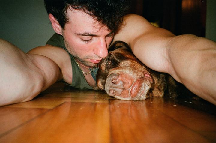 A man and a dog are lying on a wooden floor, with the man's head resting on the dog's head.