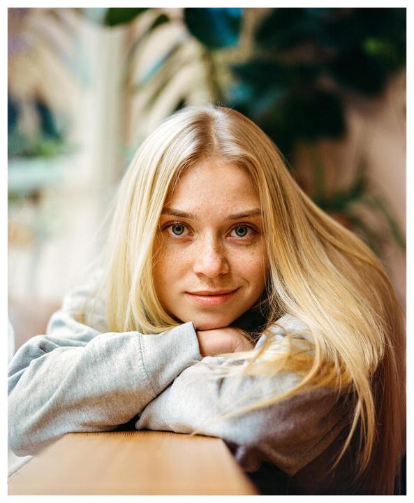 A woman with long blonde hair and freckles rests her chin on her hands, looking directly at the viewer.