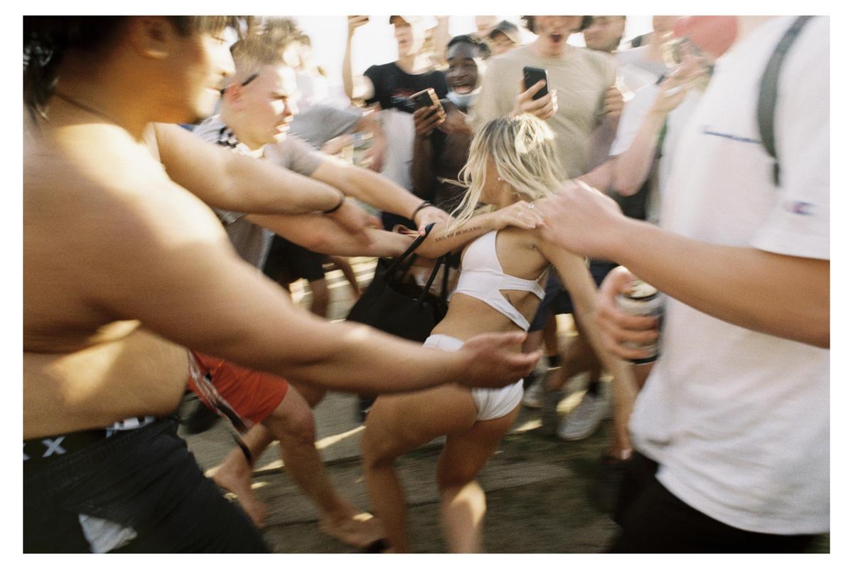 A woman in a white bikini top and shorts is surrounded by a crowd of people, some of whom are reaching out towards her.