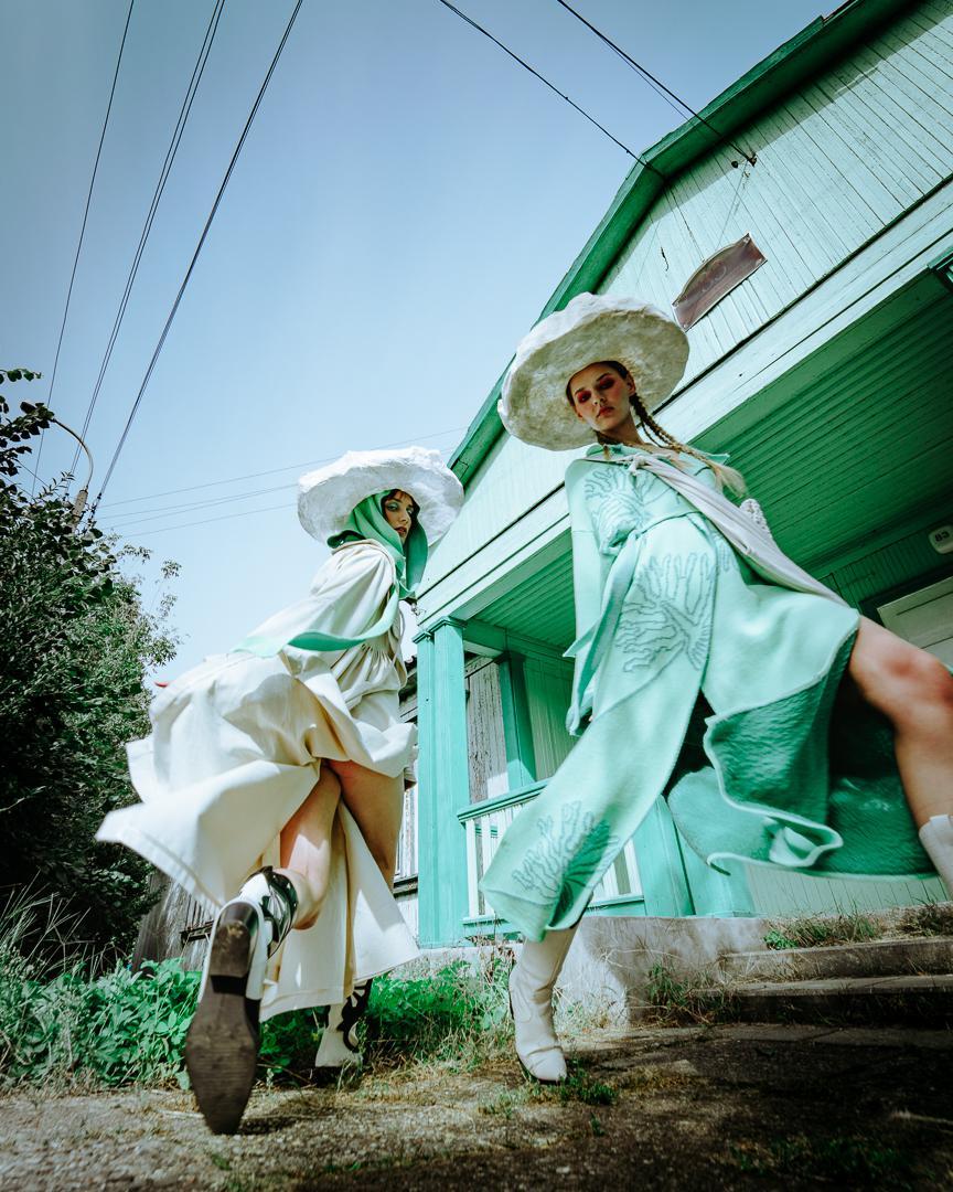 Two women in large hats and flowing dresses stand in front of a green house.