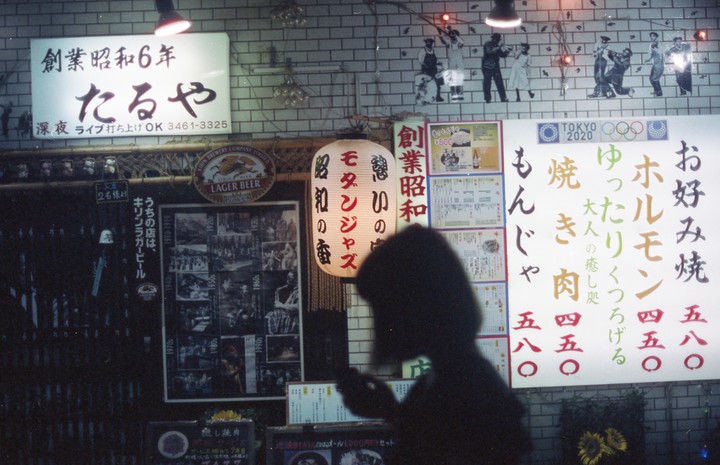 A person's silhouette is visible in front of a brightly lit Japanese restaurant sign at night.