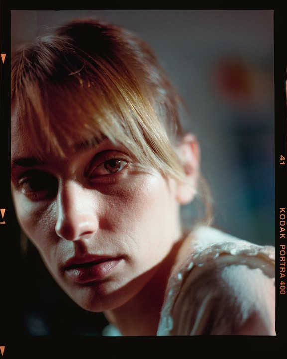 A woman with bangs looks directly at the viewer, with dramatic lighting highlighting her face.