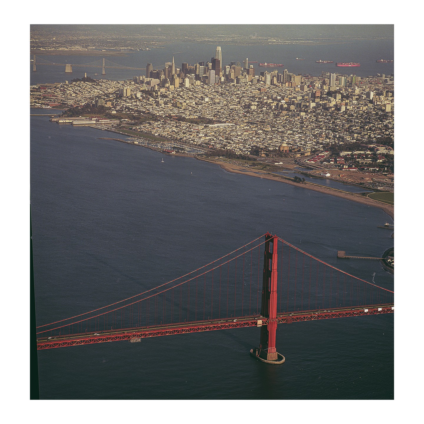An aerial view shows the Golden Gate Bridge in the foreground with the San Francisco skyline and bay in the background.