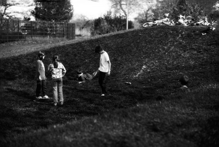 Four children play on a grassy hill with bicycles at the top.