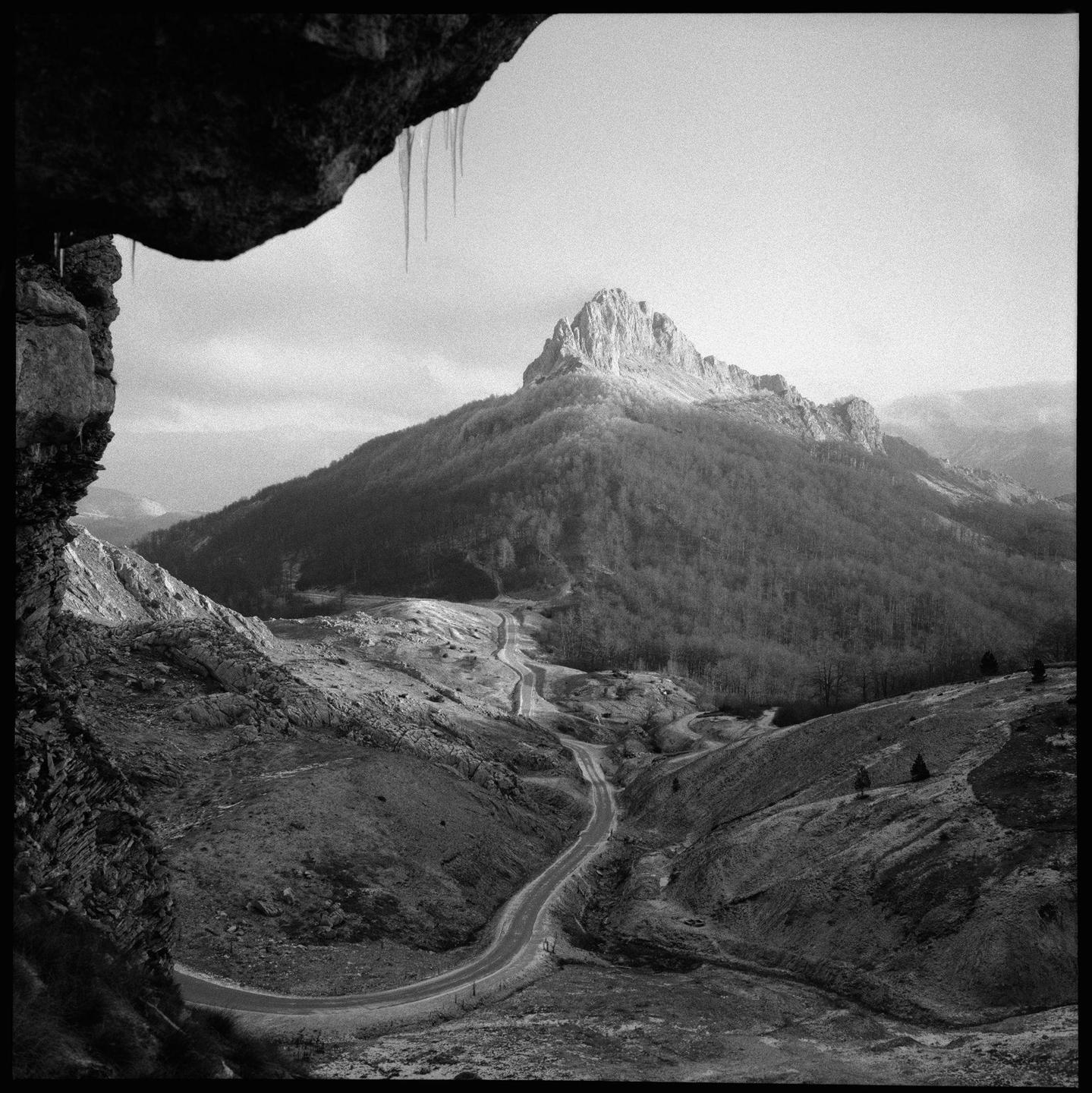 A winding road leads through a valley towards a large, rocky mountain peak under a cloudy sky, with icicles hanging from a rock formation in the foreground.