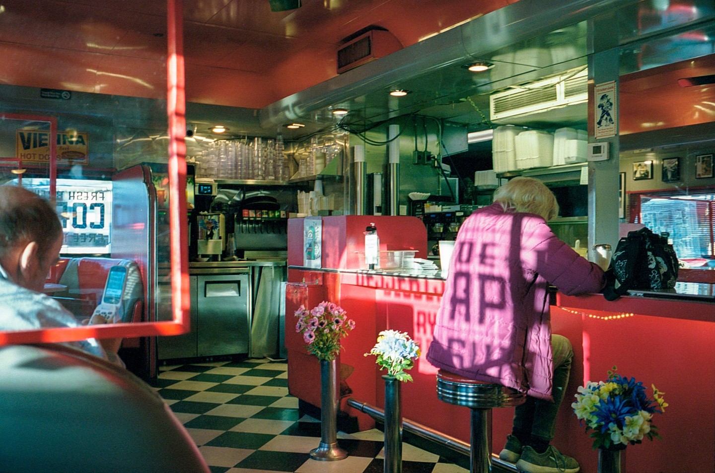 A woman sits at a red diner counter with a man in the foreground and a checkered floor.