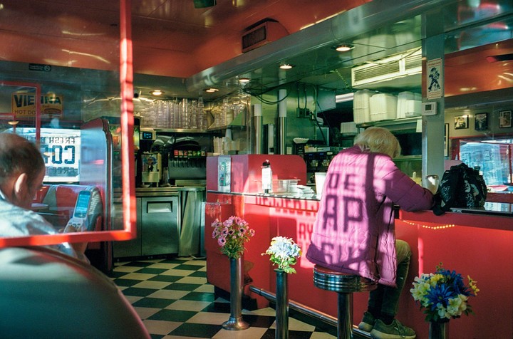 A woman sits at a red diner counter with a man in the foreground and a checkered floor.