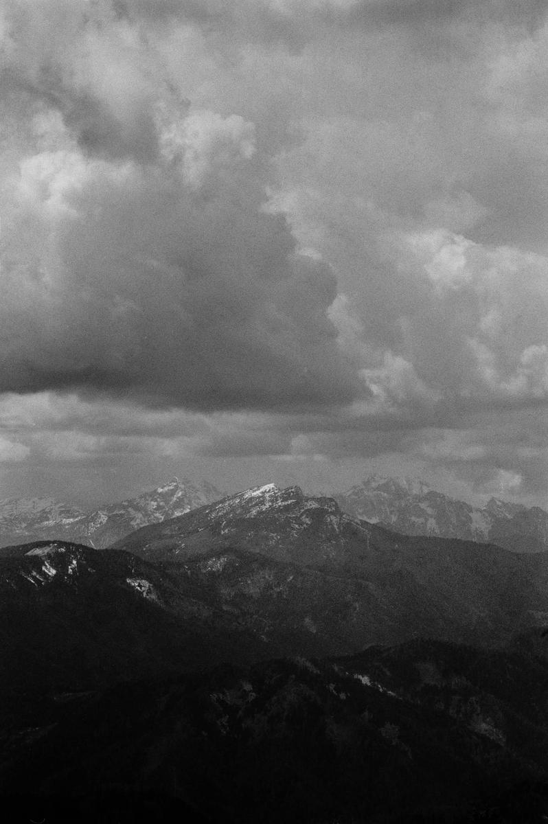 A dramatic black and white landscape shows a mountain range with snow-capped peaks under a cloudy sky.